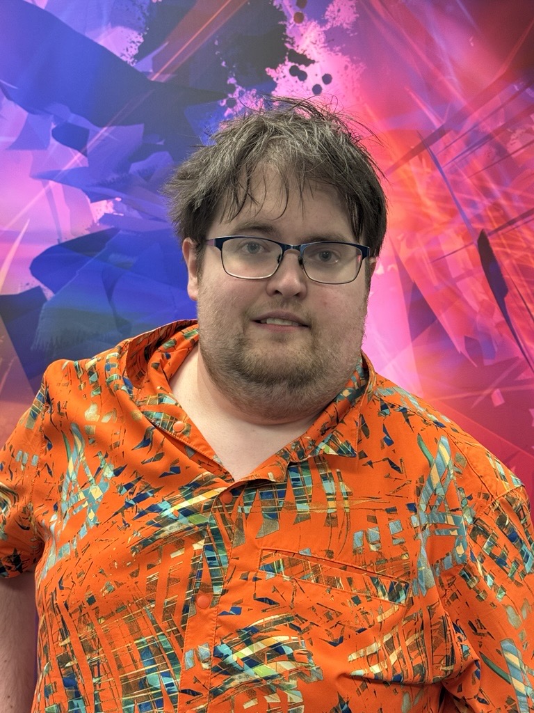 A man with short, tousled brown hair and glasses is standing in front of a colorful abstract background with shades of pink, purple, blue, and orange. He is wearing a bright orange shirt with an abstract geometric pattern in blue, green, and yellow.