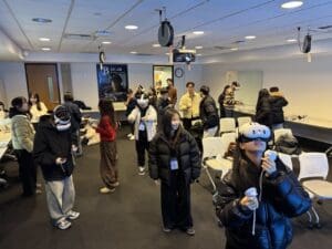 A group of students participates in a hands-on VR experience in a classroom setting. Many are wearing VR headsets and holding controllers, actively engaged in virtual activities. Others are observing, chatting, or preparing equipment. A poster on the back wall reads “XR LAB – BC Library Classroom,” indicating the event is hosted by Bellevue College’s XR Lab. The room is equipped with white chairs, projectors, and tables filled with gear, creating a lively and interactive learning environment.