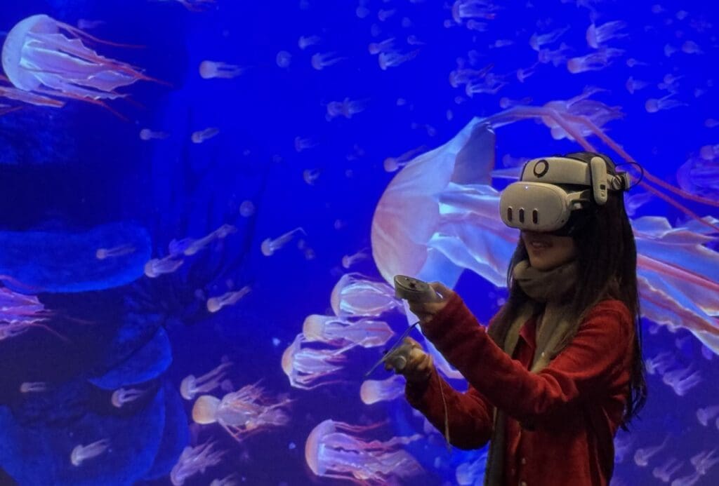 A young woman wearing a red jacket and gray scarf is immersed in a virtual reality experience featuring an underwater scene filled with glowing jellyfish. She wears a VR headset and holds motion controllers, smiling as she interacts with the digital environment. The vivid blue background with translucent jellyfish enhances the feeling of being submerged in a deep-sea world.