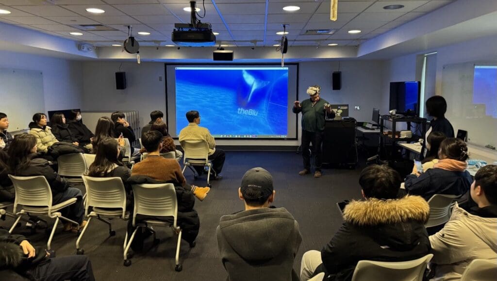A classroom at Bellevue College where students sit in a semicircle watching a VR demonstration. An instructor at the front wears a virtual reality headset and gestures with controllers, while a large projection screen behind him displays the VR experience titled "theBlu." The room is equipped with chairs, VR gear, and presentation technology, creating an immersive learning environment focused on virtual exploration.