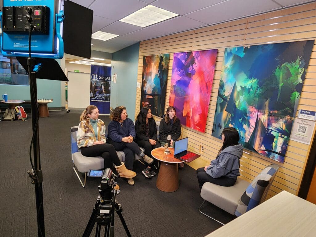 A group of five people sits in a semi-circle having a recorded discussion inside the Bellevue College XR Lab. A camera and studio light are set up in the foreground, indicating a video or podcast-style interview. A red laptop and microphone are on the table in the center. Behind the group, three large, colorful abstract art pieces hang on the wall, and a sign on the door reads “D126R XR Lab Room Booking.” A Bellevue College XR Lab banner is visible in the background.