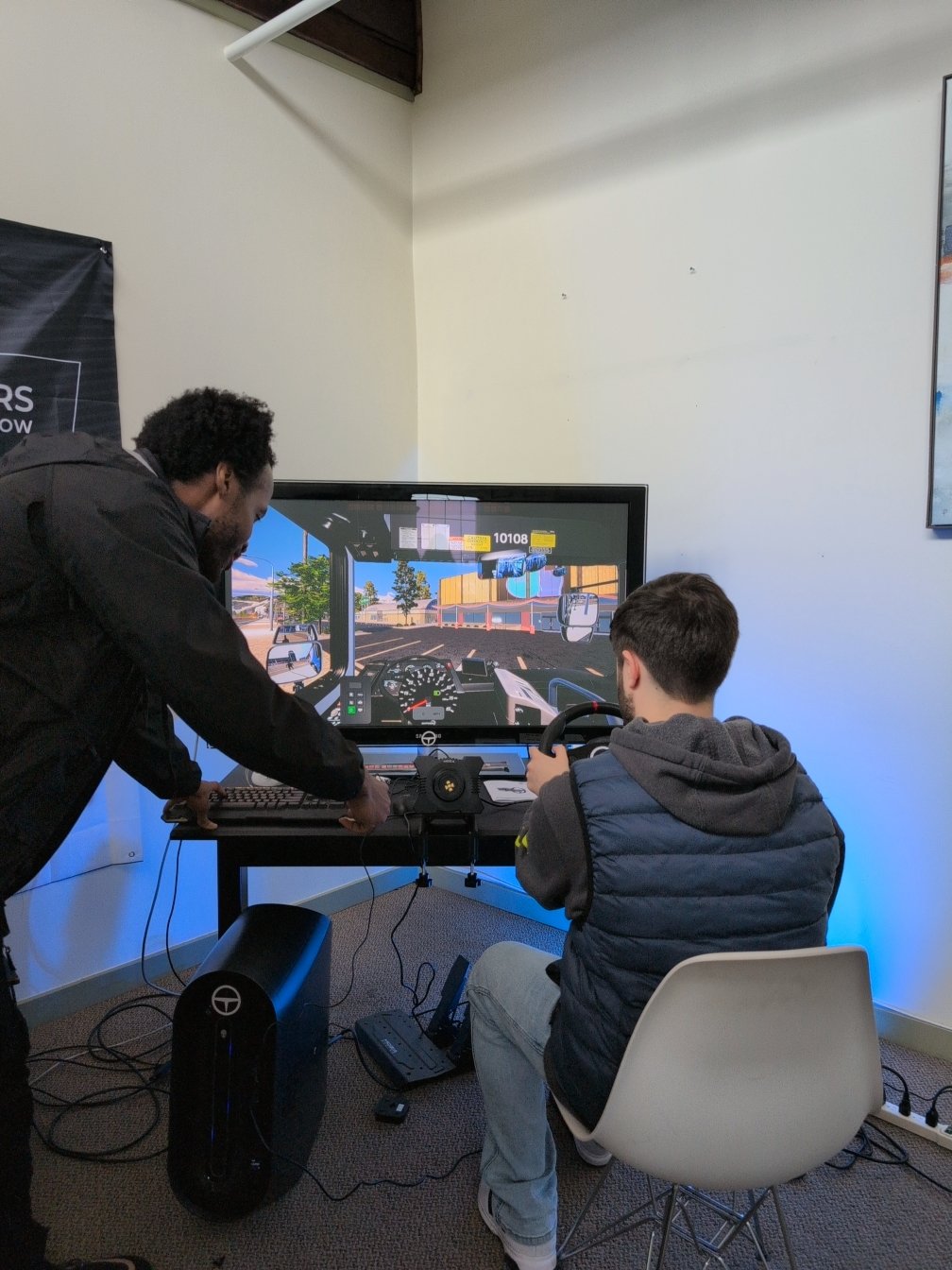 Two people are using a driving simulator setup. One person is seated in front of a monitor, holding a steering wheel and pressing pedals, while the other person is standing beside them, assisting with the keyboard and controls. The monitor displays a realistic driving simulation with a dashboard view of a truck on a road. The setup includes a desktop computer, steering wheel controller, pedals, and various cables, all placed in a room with blue accent lighting.