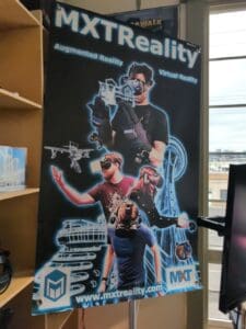 Poster for MXTReality featuring individuals wearing VR headsets and gloves, interacting with augmented and virtual reality elements such as 3D models of buildings, drones, and cruise ships. The poster includes the text "MXTReality," "Augmented Reality," "Virtual Reality," and the website "www.mxtreality.com," along with the MXTReality logo. The background shows Seattle landmarks, including the Space Needle.