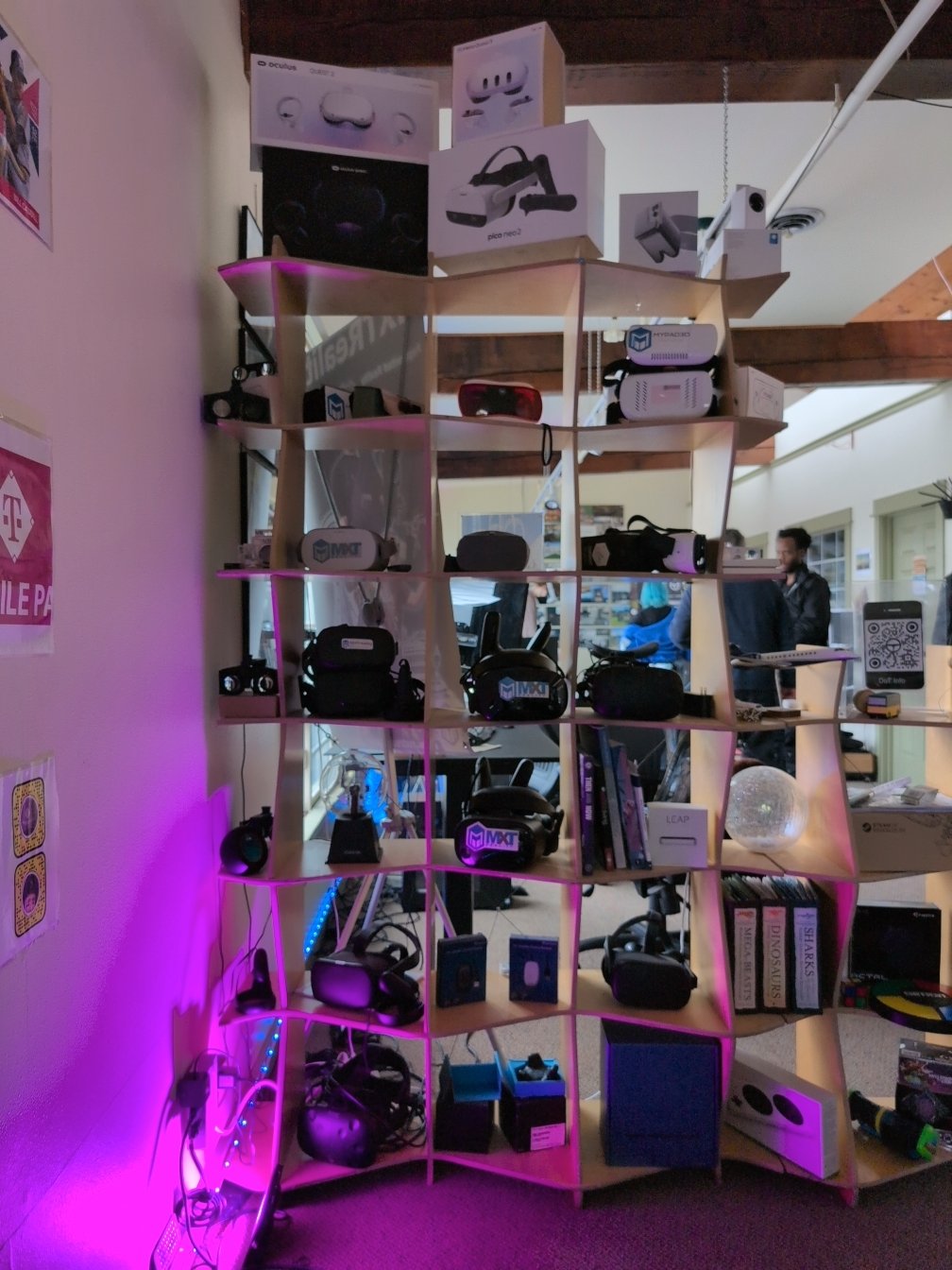 A modern, curved wooden shelf filled with various virtual reality (VR) headsets, controllers, product boxes, and educational materials. The display includes brands like Oculus, Pico, and MXTReality. Some sections also feature books, a trophy, and decorative items like a disco ball. The shelf is lit with purple accent lighting, and a workspace with people and computers is visible in the background.