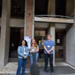 Three Bellevue College students stand in front of the Meany Studio Theatre entrance at the University of Washington. They are smiling and dressed casually, with a guitar case positioned in front of the student in the center. Behind them is a sign that reads “Welcome – Performing Arts Session” as part of the Undergraduate Research Symposium.