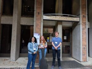 Three Bellevue College students stand in front of the Meany Studio Theatre entrance at the University of Washington. They are smiling and dressed casually, with a guitar case positioned in front of the student in the center. Behind them is a sign that reads “Welcome – Performing Arts Session” as part of the Undergraduate Research Symposium.