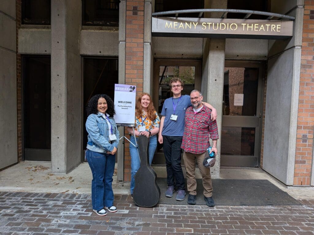 Four people stand smiling outside the entrance to Meany Studio Theatre at the University of Washington. A sign next to them reads “Welcome – Performing Arts Session” as part of the Undergraduate Research Symposium. The group includes three Bellevue College students and an instructor, with one holding a guitar case, indicating their participation in a performance-based research project.