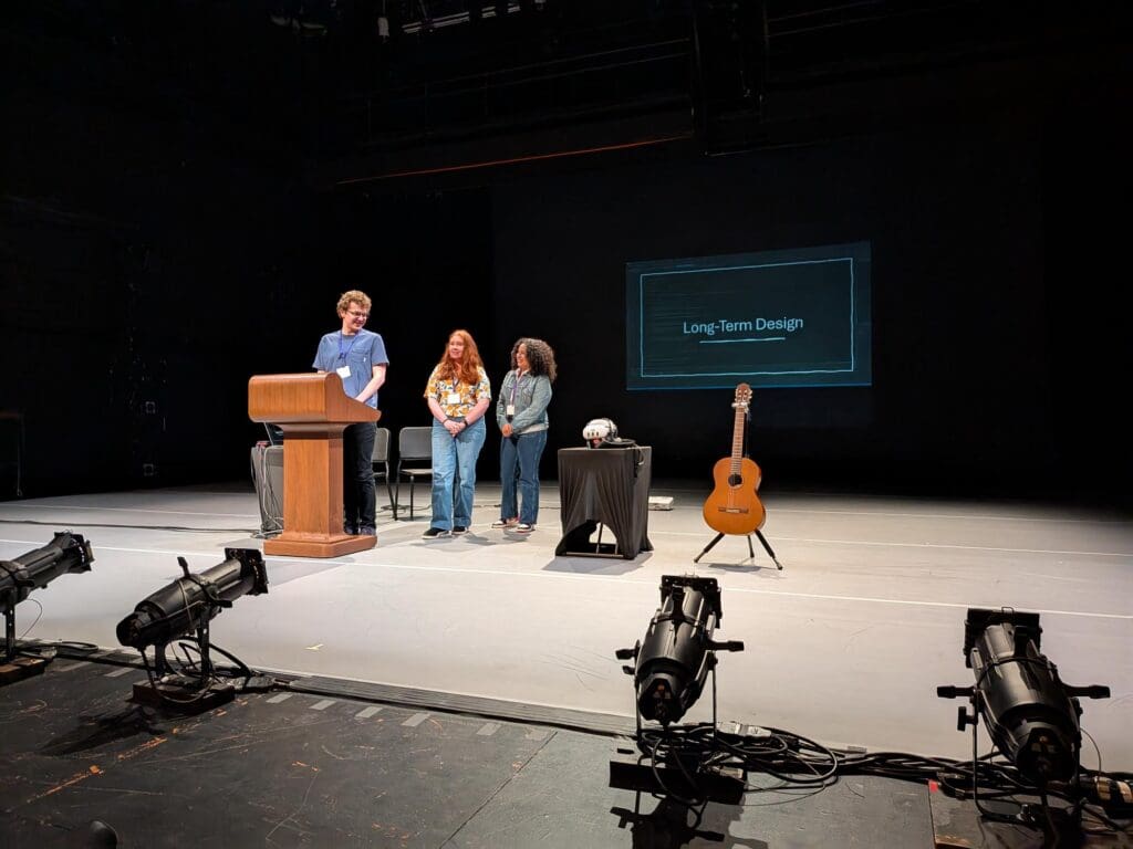 Three Bellevue College students present their project on stage at Meany Studio Theatre during the UW Undergraduate Research Symposium. One student speaks at a podium while the other two stand behind him, smiling. A slide projected behind them reads “Long-Term Design.” Nearby, a classical guitar and a VR headset sit on a table, reflecting the blend of music and immersive technology in their project. Stage lighting equipment is visible at the front of the scene.