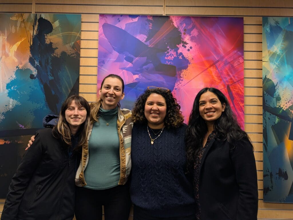 Four women stand smiling side by side in front of a colorful abstract art wall display inside the Bellevue College XR Lab. They appear to be participants or collaborators in a project or event, showing a strong sense of camaraderie. The background features three large, vibrant panels with layered digital art in shades of blue, purple, orange, and pink.
