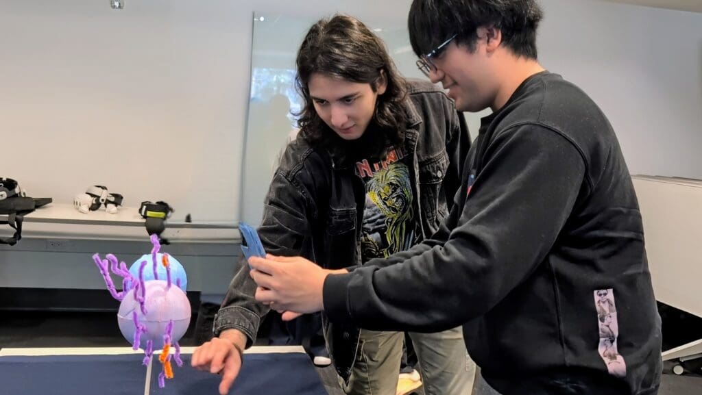 Two students at Bellevue College closely examine a colorful handmade anatomical model made from foam spheres and pipe cleaners, capturing it with a smartphone. They appear focused as they prepare the model for 3D scanning. In the background, VR headsets rest on a nearby table, indicating the activity is part of an XR Lab project that combines physical modeling with digital tools.