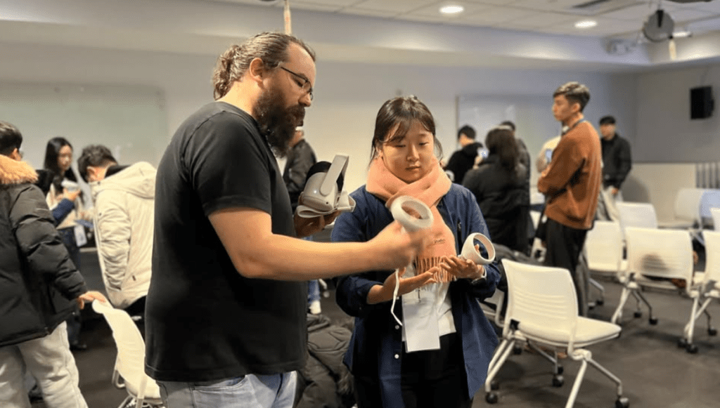 In a classroom setting, a man with glasses and a beard is demonstrating how to use a VR headset and controllers to a young woman wearing a pink scarf. The woman attentively examines the controllers in her hands. Other students in the background are either preparing equipment or engaged in conversation, suggesting a hands-on VR workshop or training session. White chairs and modern classroom equipment fill the room.