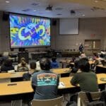 A large group of people seated in a lecture hall watch a colorful presentation on a big screen at the front of the room. The screen displays the words "Global Game Jam 2025" with vibrant graphics. Attendees appear to be participants or guests of the event, many with laptops, notebooks, or plates of food. The room is equipped with projectors, presentation equipment, and catering tables along the side wall.