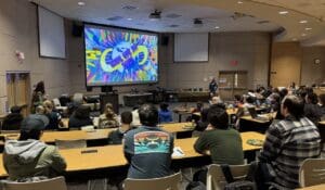 A large group of people seated in a lecture hall watch a colorful presentation on a big screen at the front of the room. The screen displays the words "Global Game Jam 2025" with vibrant graphics. Attendees appear to be participants or guests of the event, many with laptops, notebooks, or plates of food. The room is equipped with projectors, presentation equipment, and catering tables along the side wall.