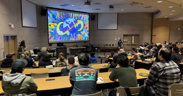 A large group of people at the 2025 Global Jam seated in a lecture hall watch a colorful presentation on a big screen that has vibrant graphics.