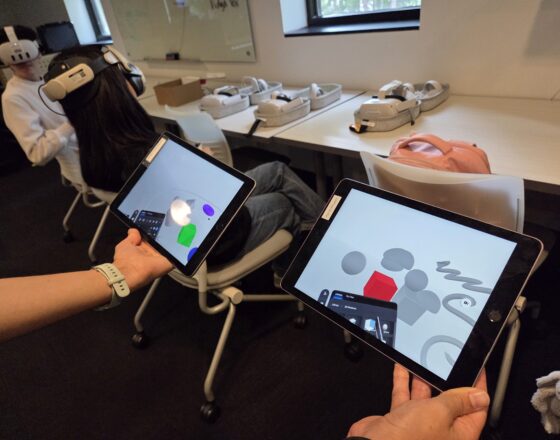 Hands holding two ipads showing two version of an XR painting application. People with headsets sit in background.